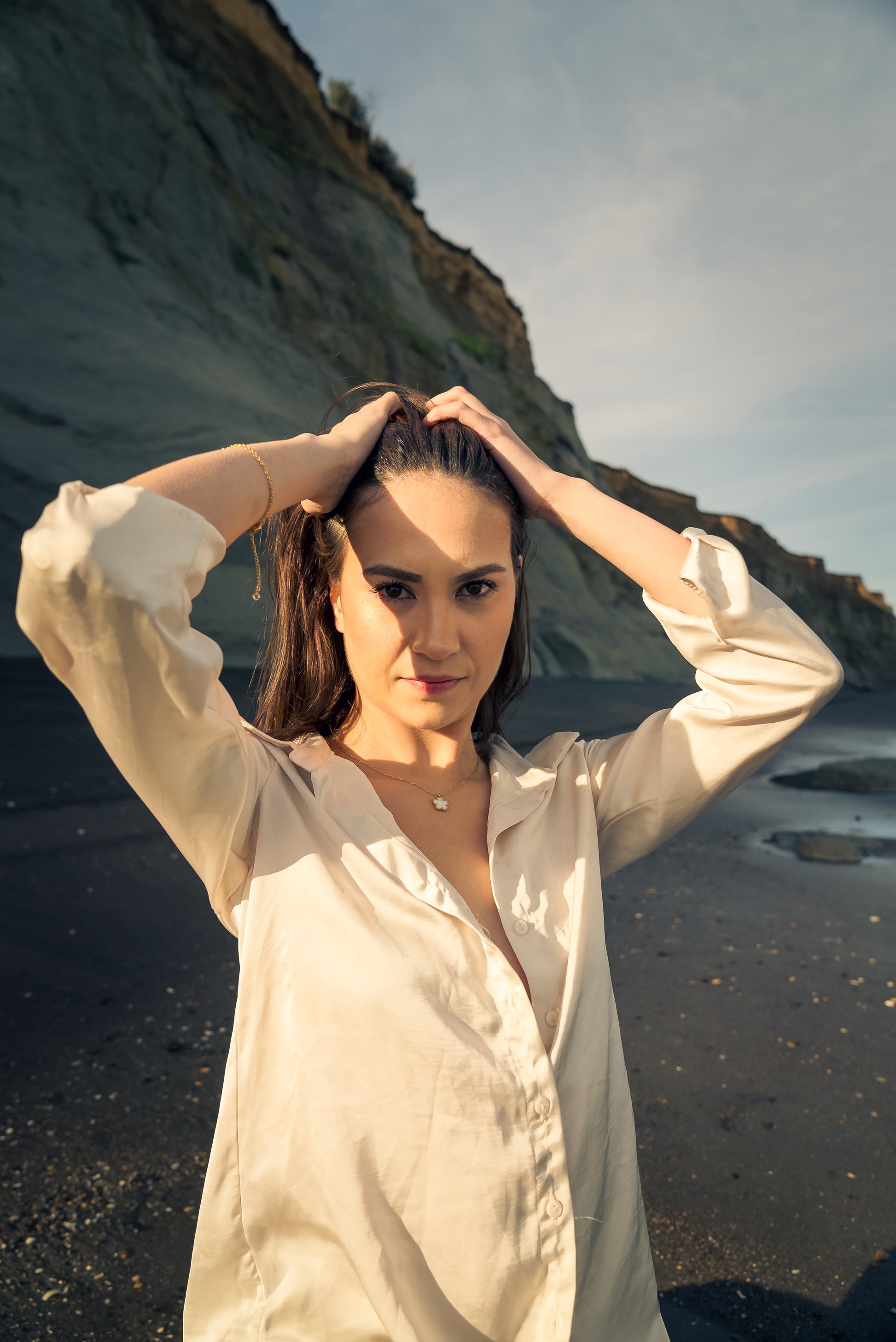 Woman wearing gold Lily Necklace  adjusting her hair on a beach with cliffs in the background