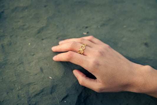 Hand wearing a gold ring with interlocked oval rings on a textured surface. The ring size is adjustable.